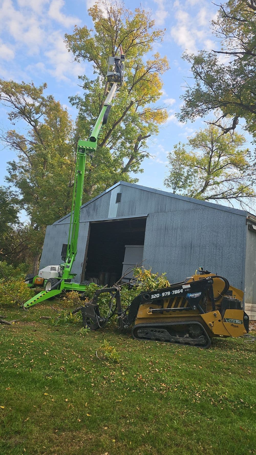High reach lift performing tree trimming near a barn with landscaping equipment nearby.