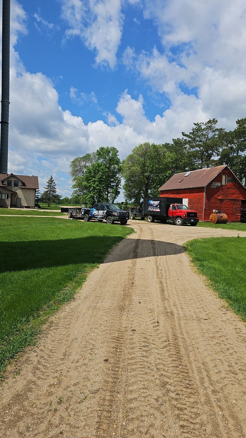 Gravel road leading to a red barn and trucks on a sunny farm landscape with blue skies.