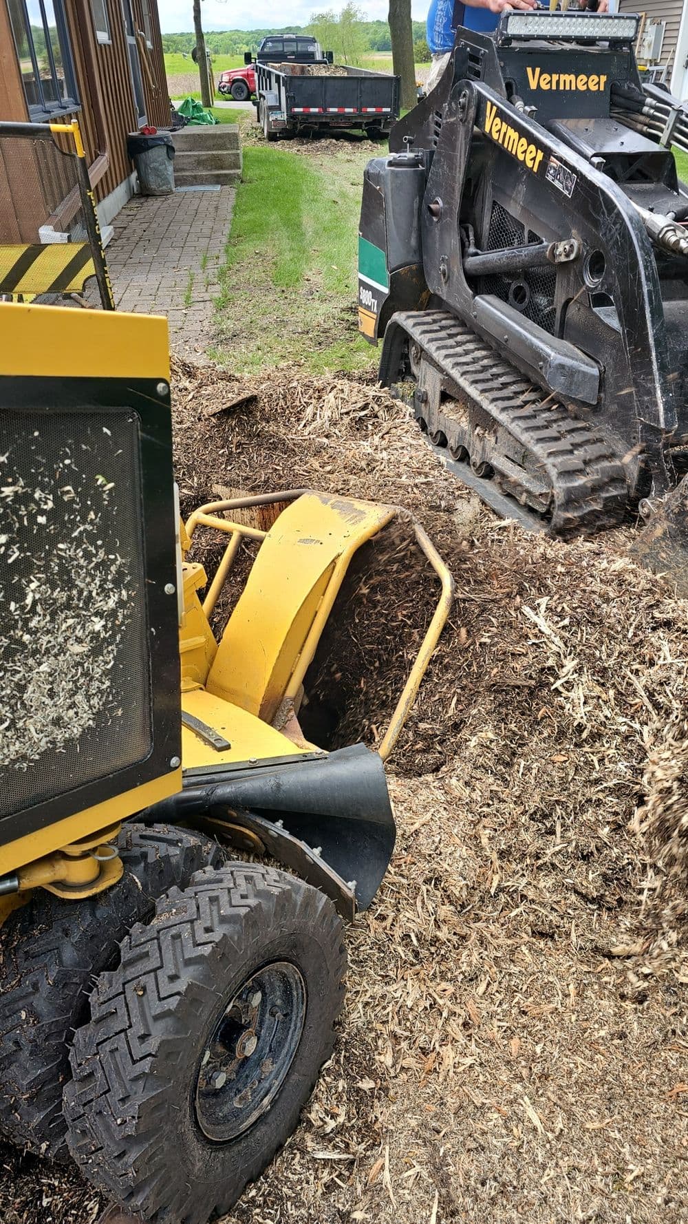 Stump grinder and compact track loader on mulch-covered ground at a landscaping site.