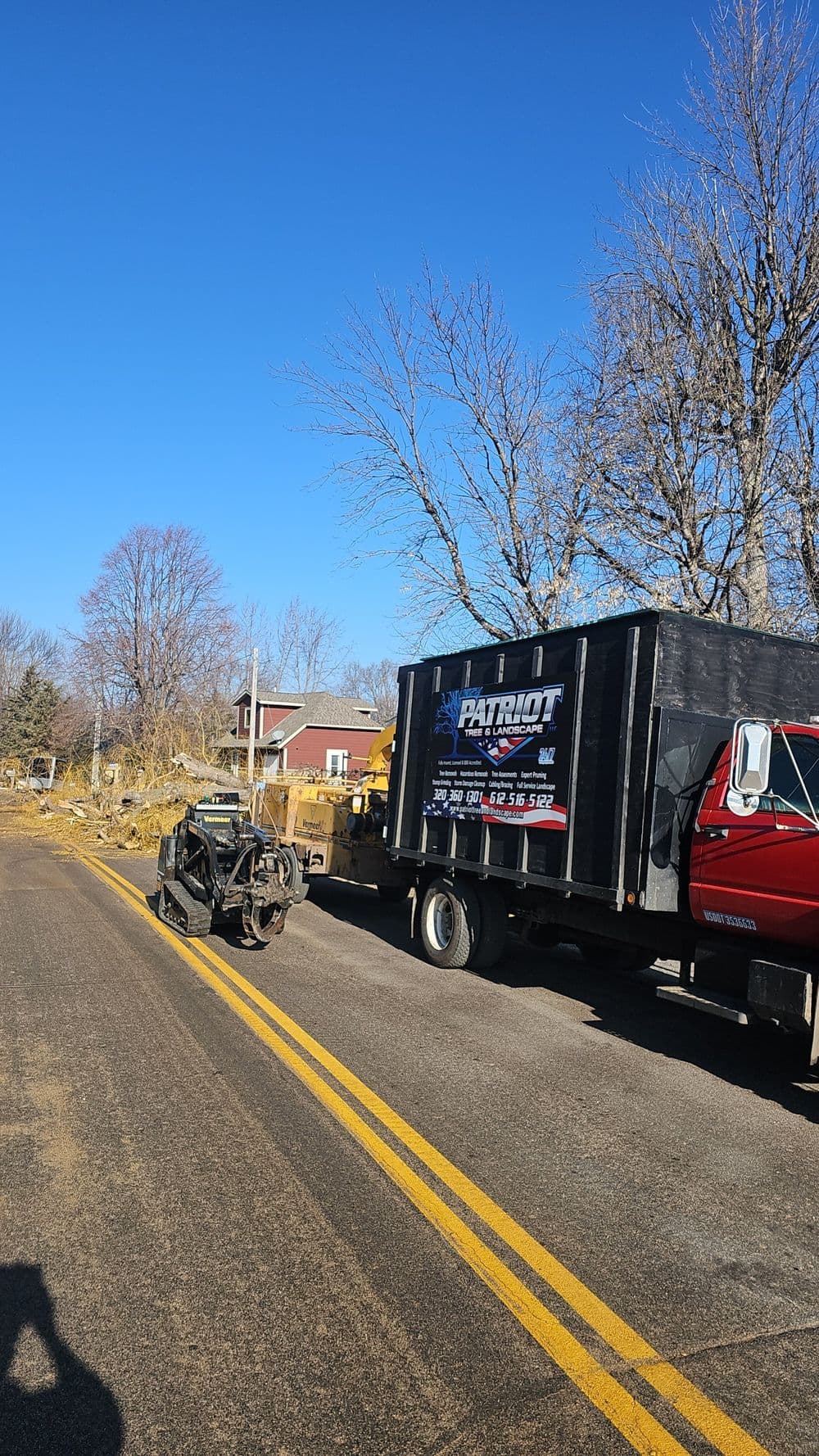 Tow truck with disabled vehicle on rural road under clear blue sky. Nearby trees and houses visible.