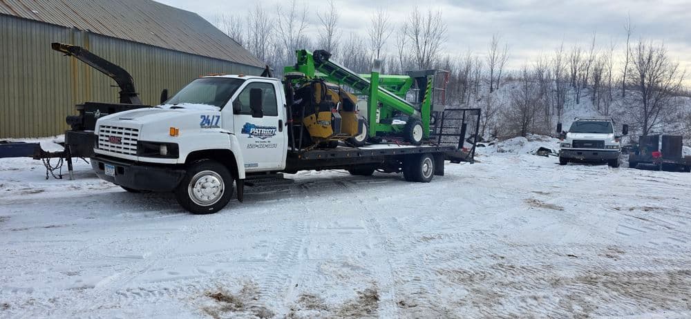 Flatbed truck transporting green equipment in a snowy yard with industrial backdrop.