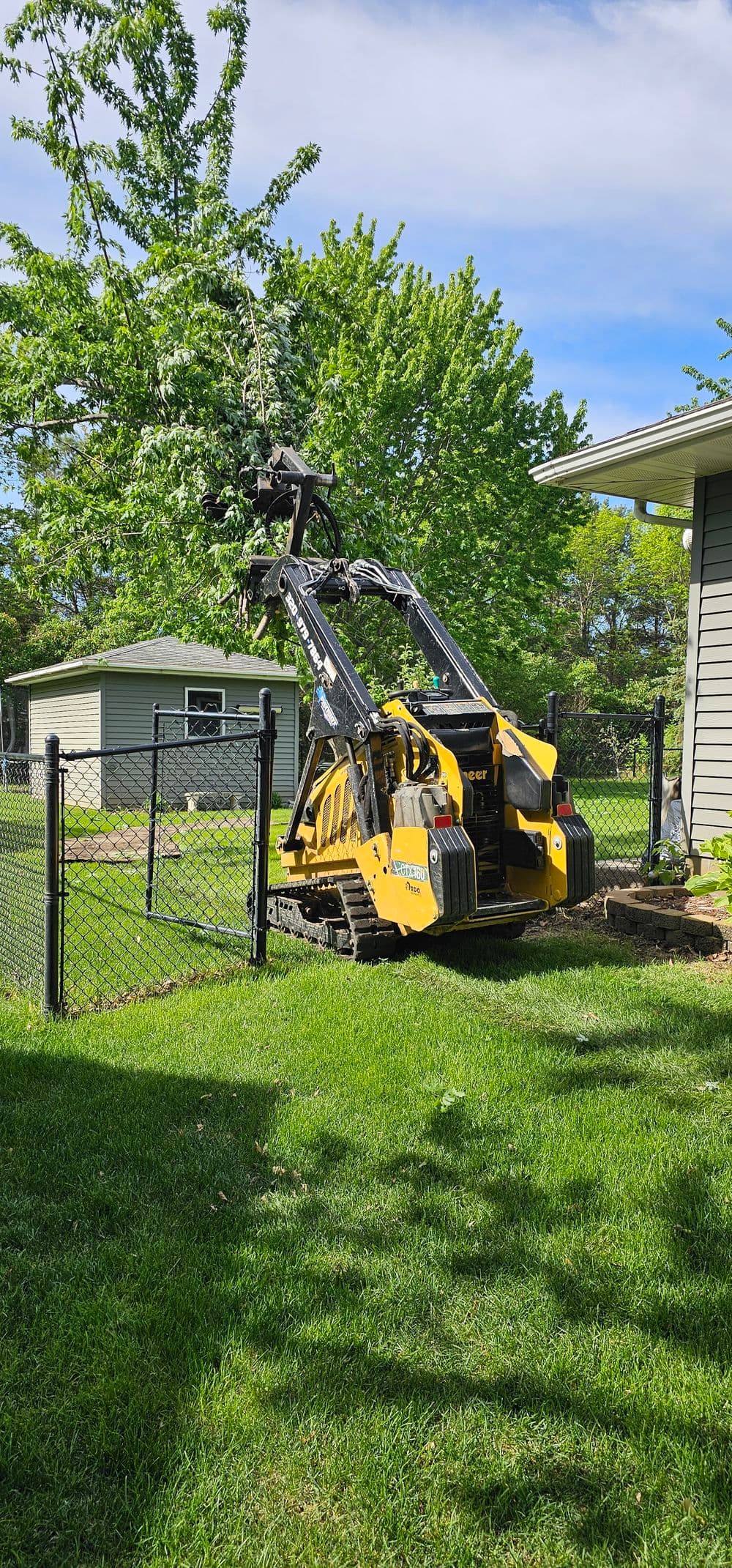 Compact track loader behind a fence, working in a lush green yard with trees.