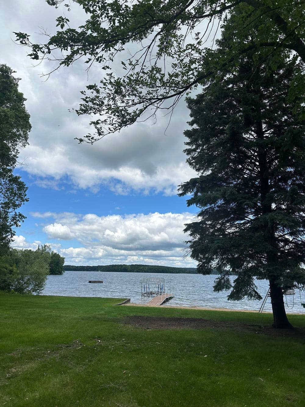Scenic lake view with cloudy sky, green grass, and a dock surrounded by trees.