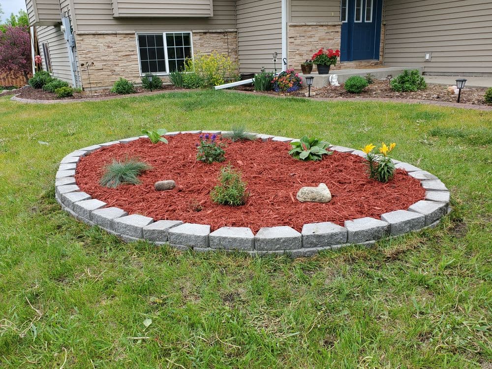 Circular flower bed with mulch and various plants, surrounded by a stone border.