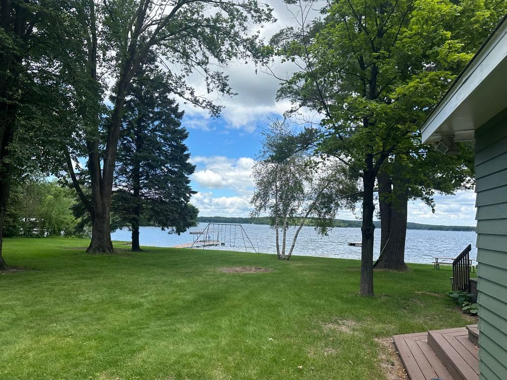 Scenic lakeside view with green grass, trees, and a swing set against a cloudy sky.