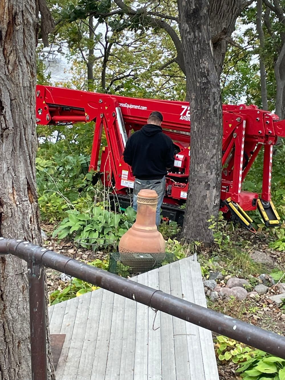Man operating a red lift near a lake, surrounded by trees and greenery.