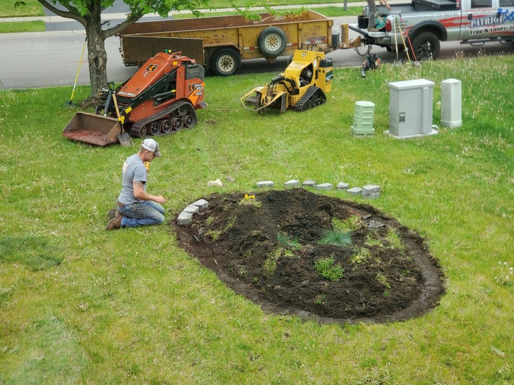 Landscape gardener planting in an oval garden bed with machinery and tools nearby.