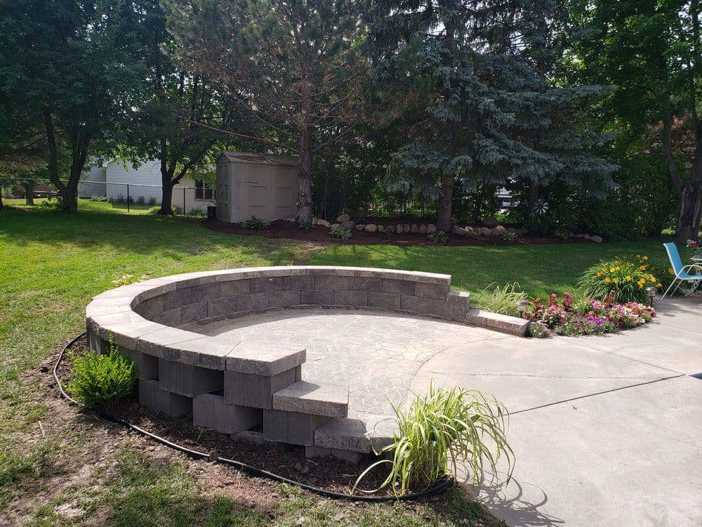 Curved stone patio surrounded by lush green grass and colorful flowers in a tranquil backyard.