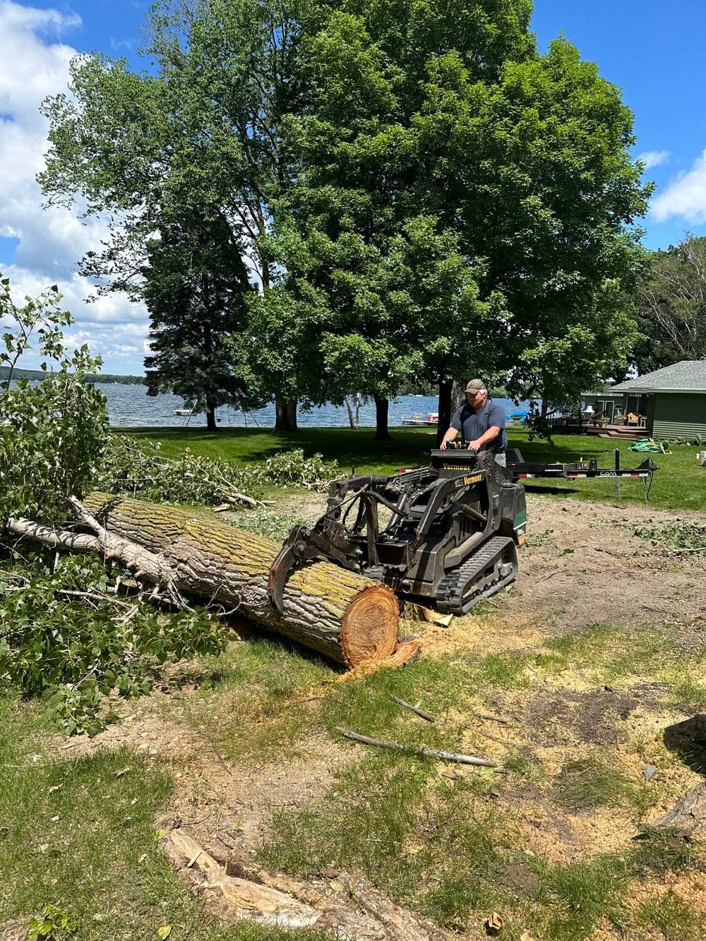 Man operating a skid steer to move logs in a lakeside yard with greenery and a house in the background.