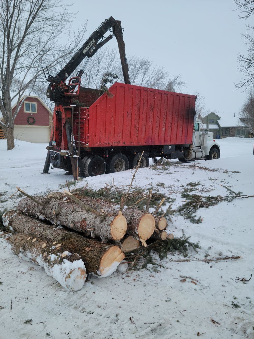 Logging truck with crane loading felled trees in snowy residential area.