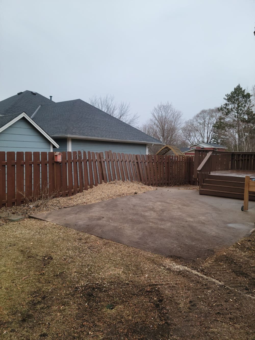 Empty backyard patio with wooden fence and gray house in the background, overcast sky.