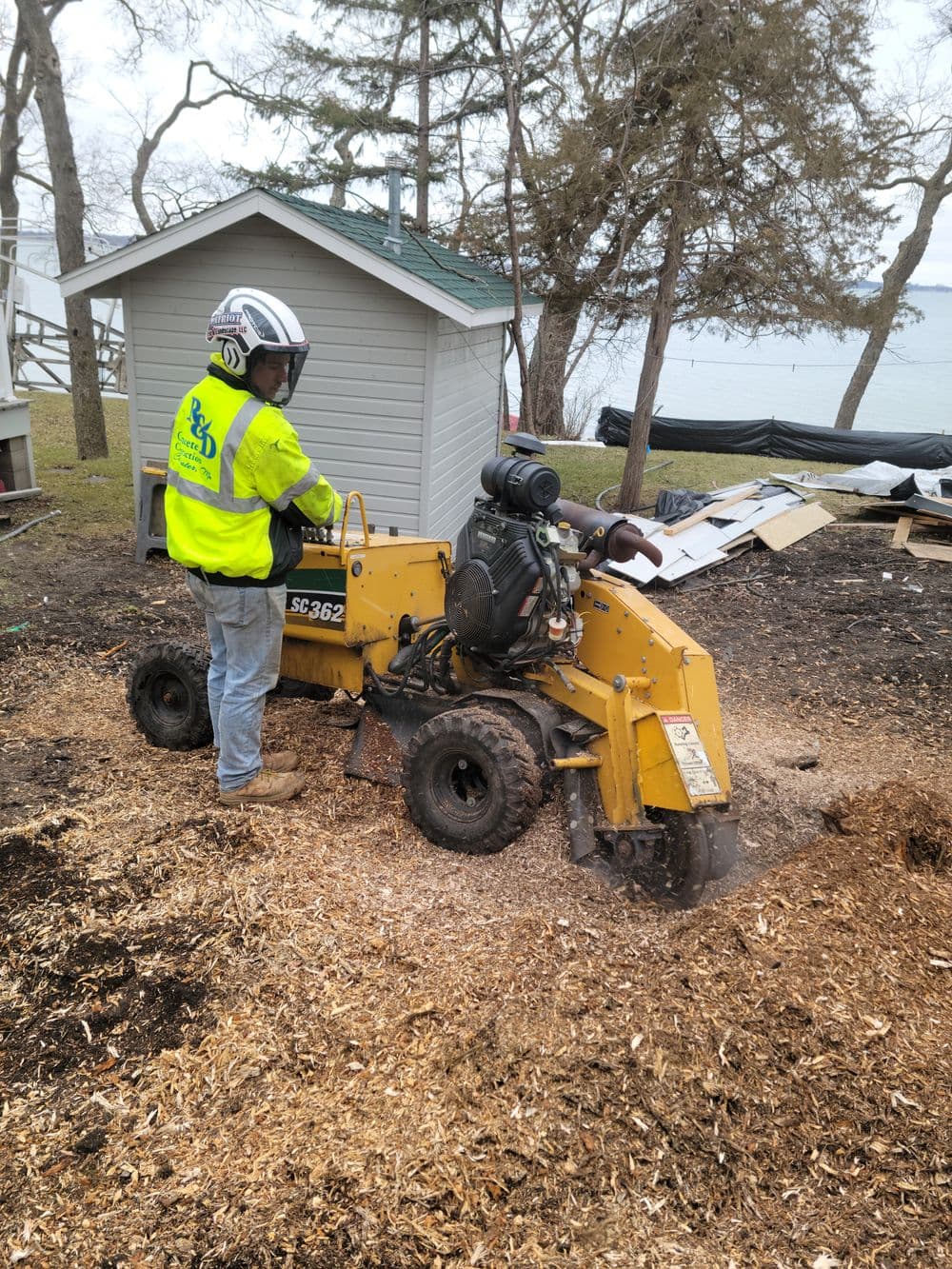Tree stump grinder in action near a lake, with operator in safety gear and yellow machinery.