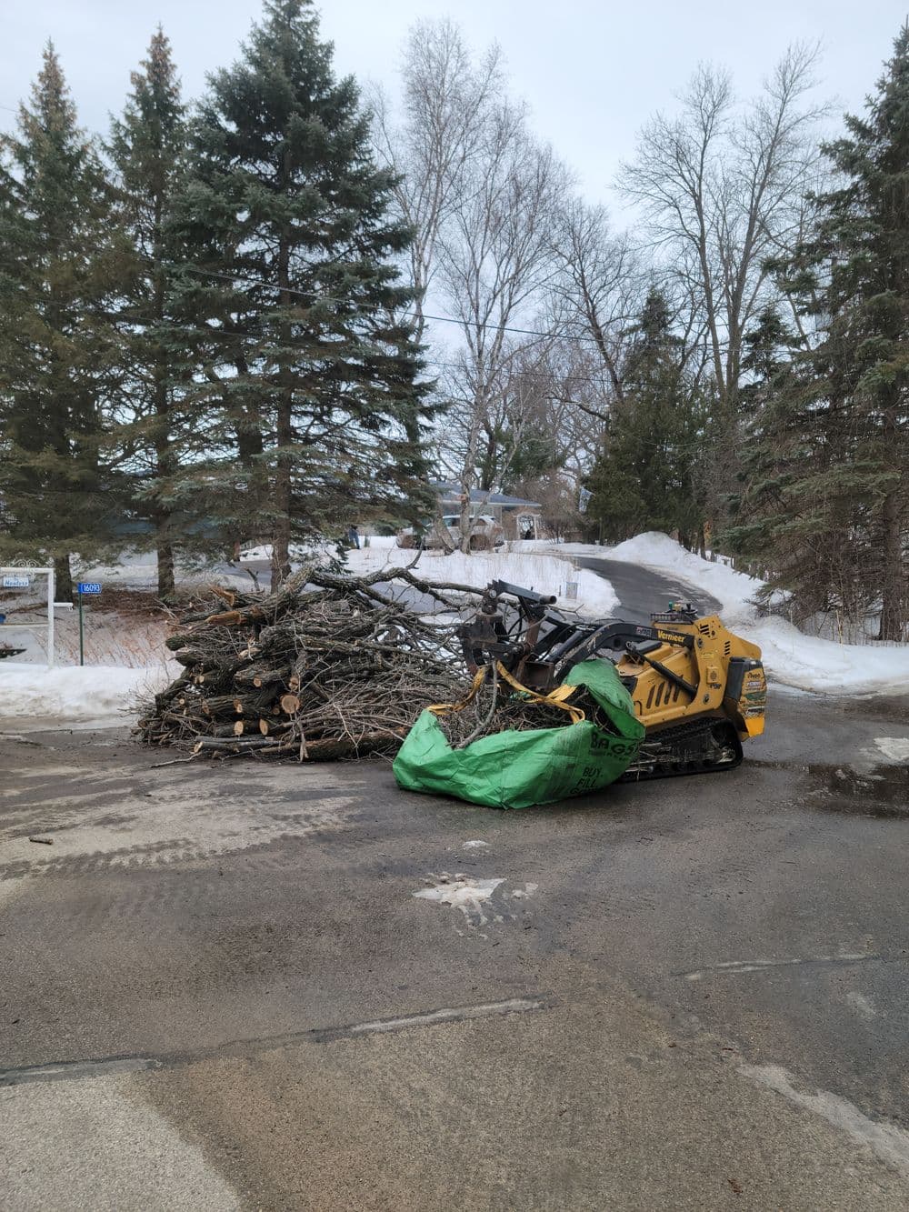 Skid steer loader clearing winter debris in a residential area with stacked branches.