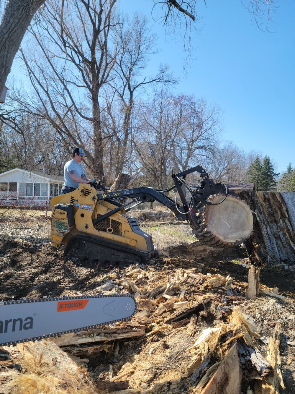Person operating a stump grinder to remove a tree stump in a sunny yard.